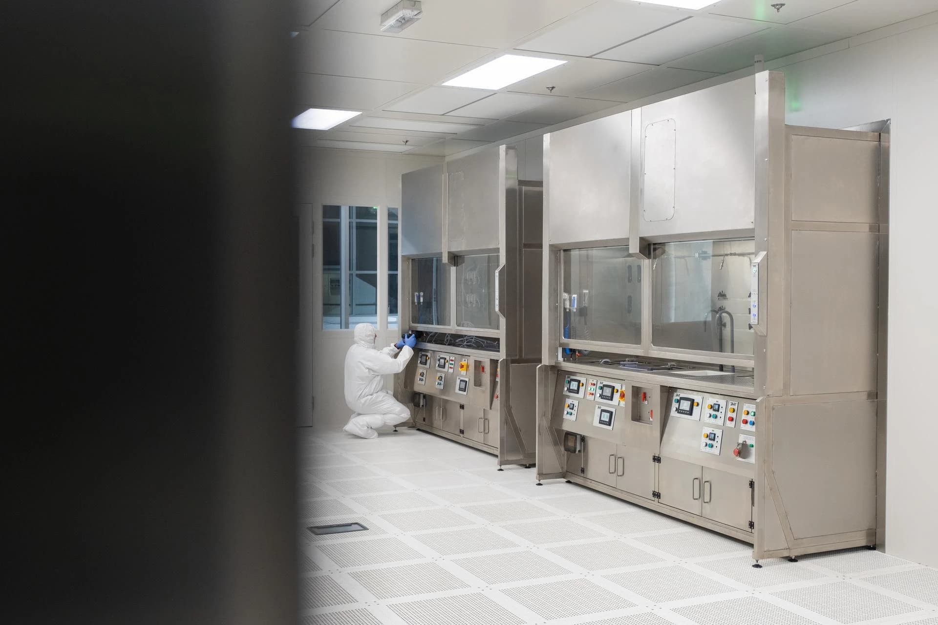 A person in a white cleanroom suit and blue gloves crouches while working at a stainless steel workstation equipped with control screens and buttons. Two such modern stainless steel workstations are present in the brightly lit, sterile cleanroom with a white perforated raised floor.