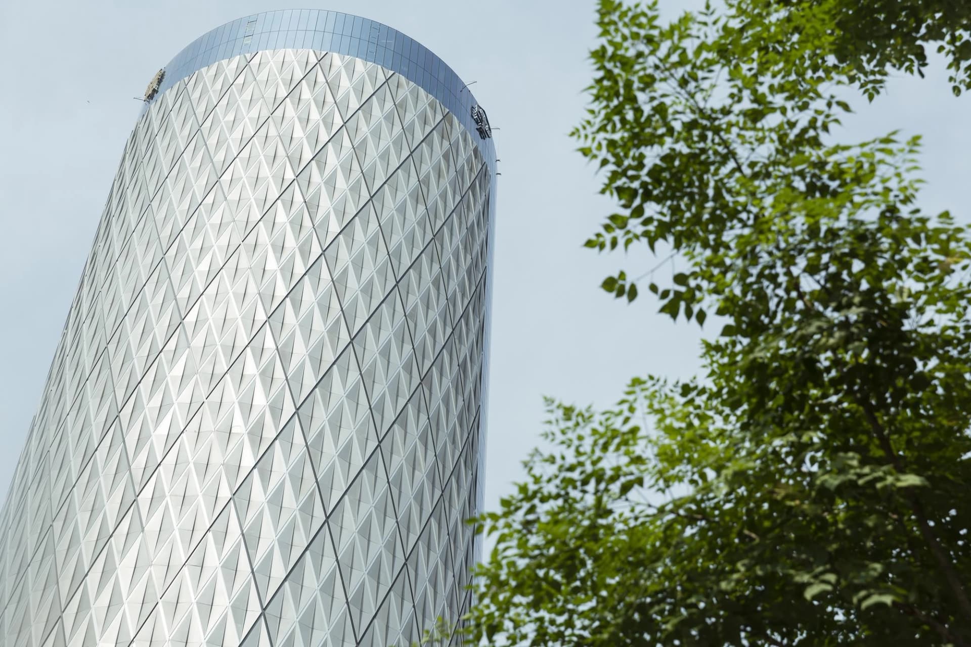 Gratte-ciel moderne à façade en losanges s'élevant derrière des feuilles d'arbres vertes sous un ciel bleu clair.