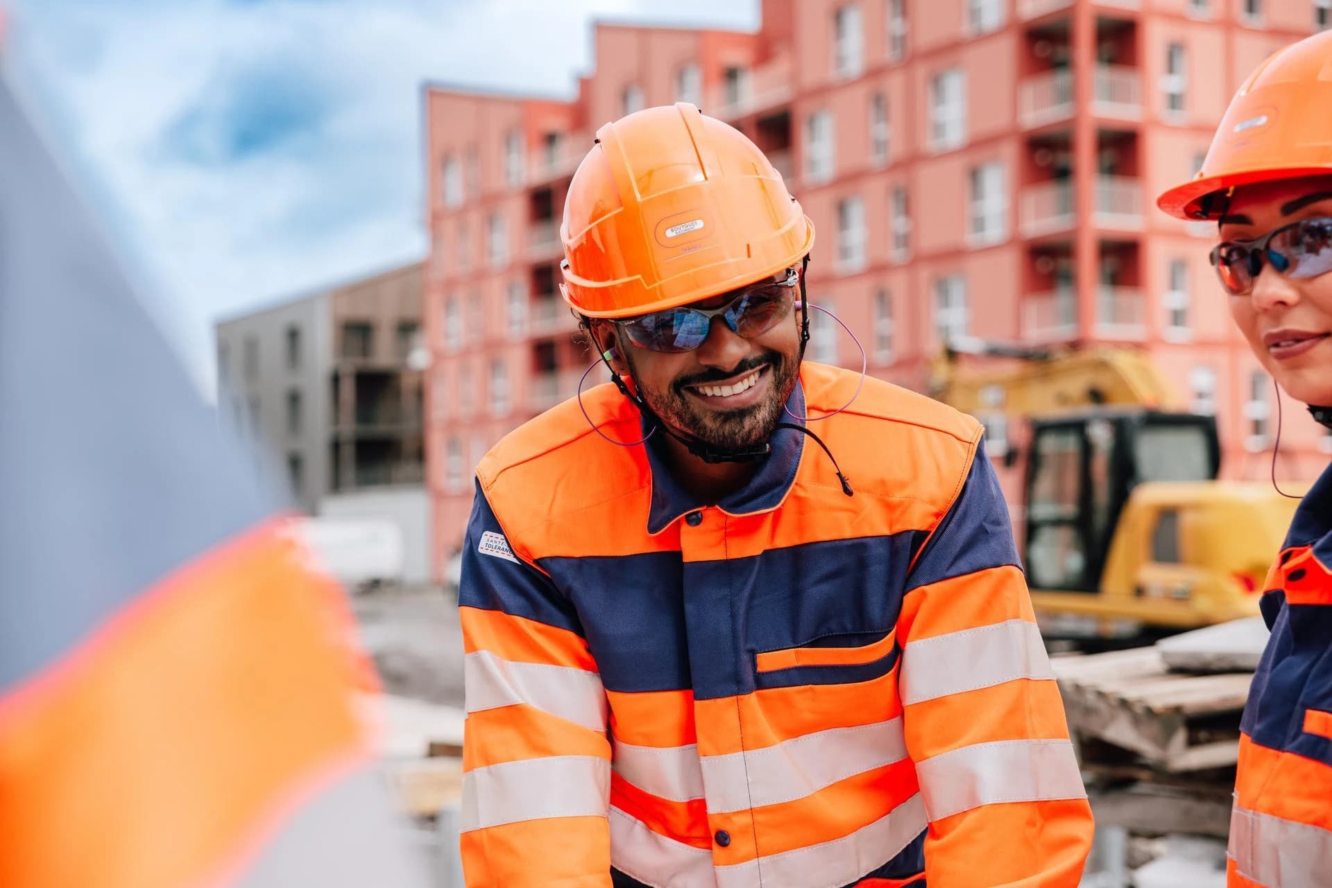 Gros plan sur un homme souriant portant un casque de sécurité orange, des lunettes et une veste de chantier orange et bleue à bandes réfléchissantes. En arrière-plan flou, un site de construction avec des bâtiments roses et un autre travailleur sont visibles.