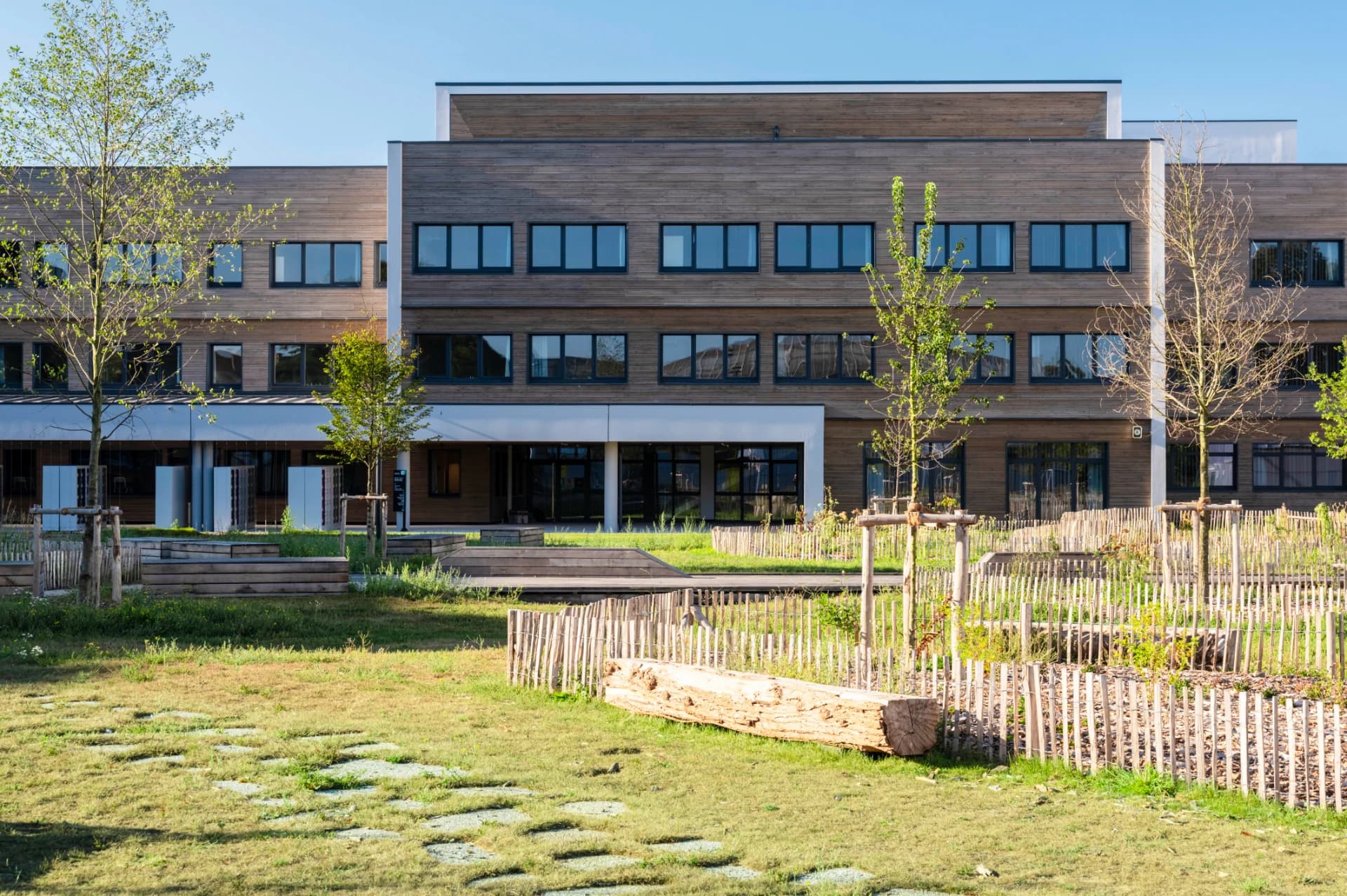 Bâtiment moderne de trois étages à la façade en bois clair et aux nombreuses fenêtres sombres, avec une pelouse et un jardin paysager de jeunes arbres, de clôtures basses et d'une bûche au premier plan, sous un ciel bleu.