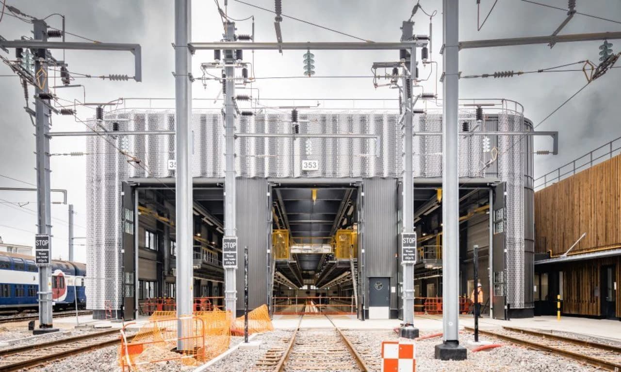 Vue extérieure d'un dépôt de maintenance ferroviaire moderne avec des voies ferrées menant à un grand bâtiment industriel à façade métallique, sous un ciel nuageux.