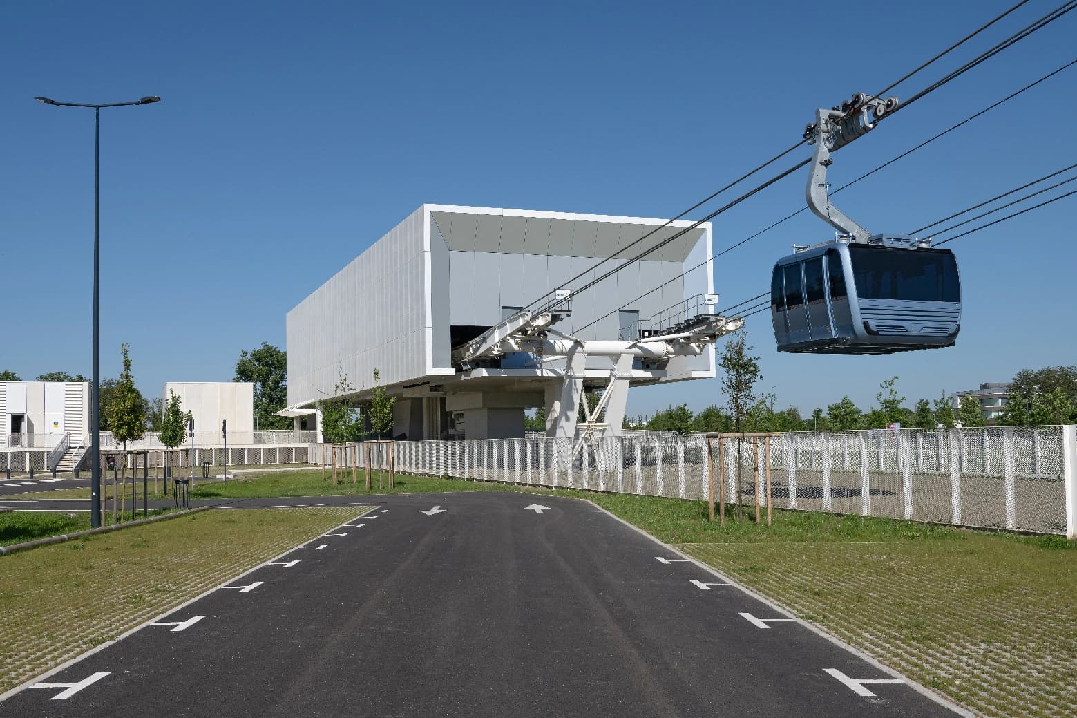 Gare de téléphérique moderne et blanche avec une cabine grise suspendue sous un ciel bleu clair. Une allée goudronnée mène vers la station, bordée de verdure.