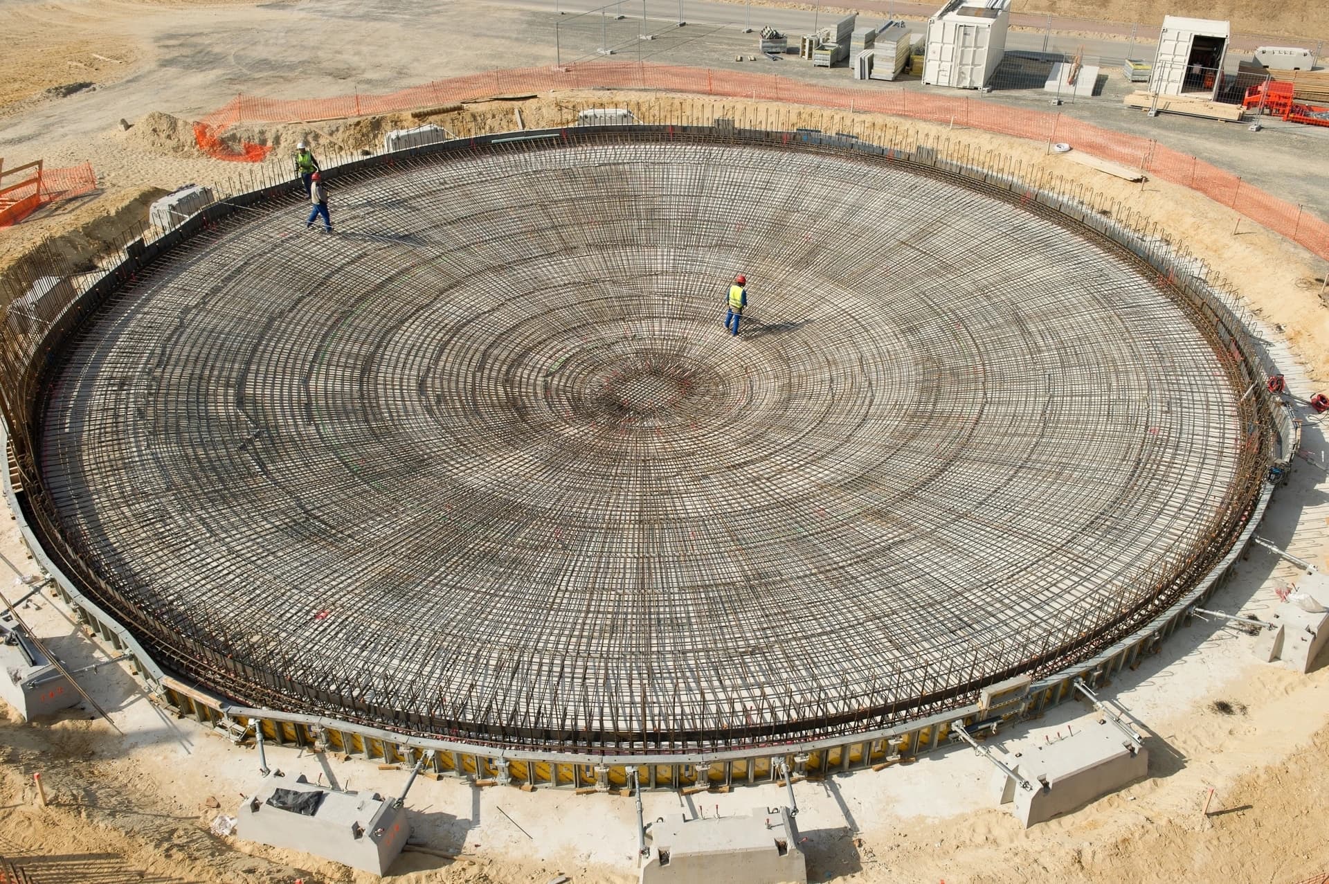 Vue aérienne d'une immense fondation circulaire en béton armé, montrant un dense treillis de barres d'armature métalliques où travaillent des ouvriers en gilets jaunes. Le chantier sablonneux est entouré de clôtures de sécurité orange.