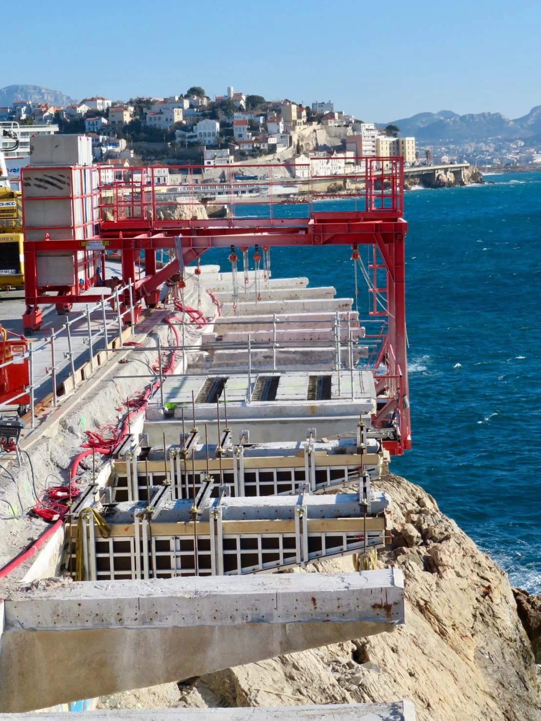 Chantier de construction sur une falaise rocheuse, avec une structure de support rouge surplombant une mer d'un bleu profond. Des éléments en béton sont en cours de montage, et une ville côtière s'étend en arrière-plan sous un ciel clair.