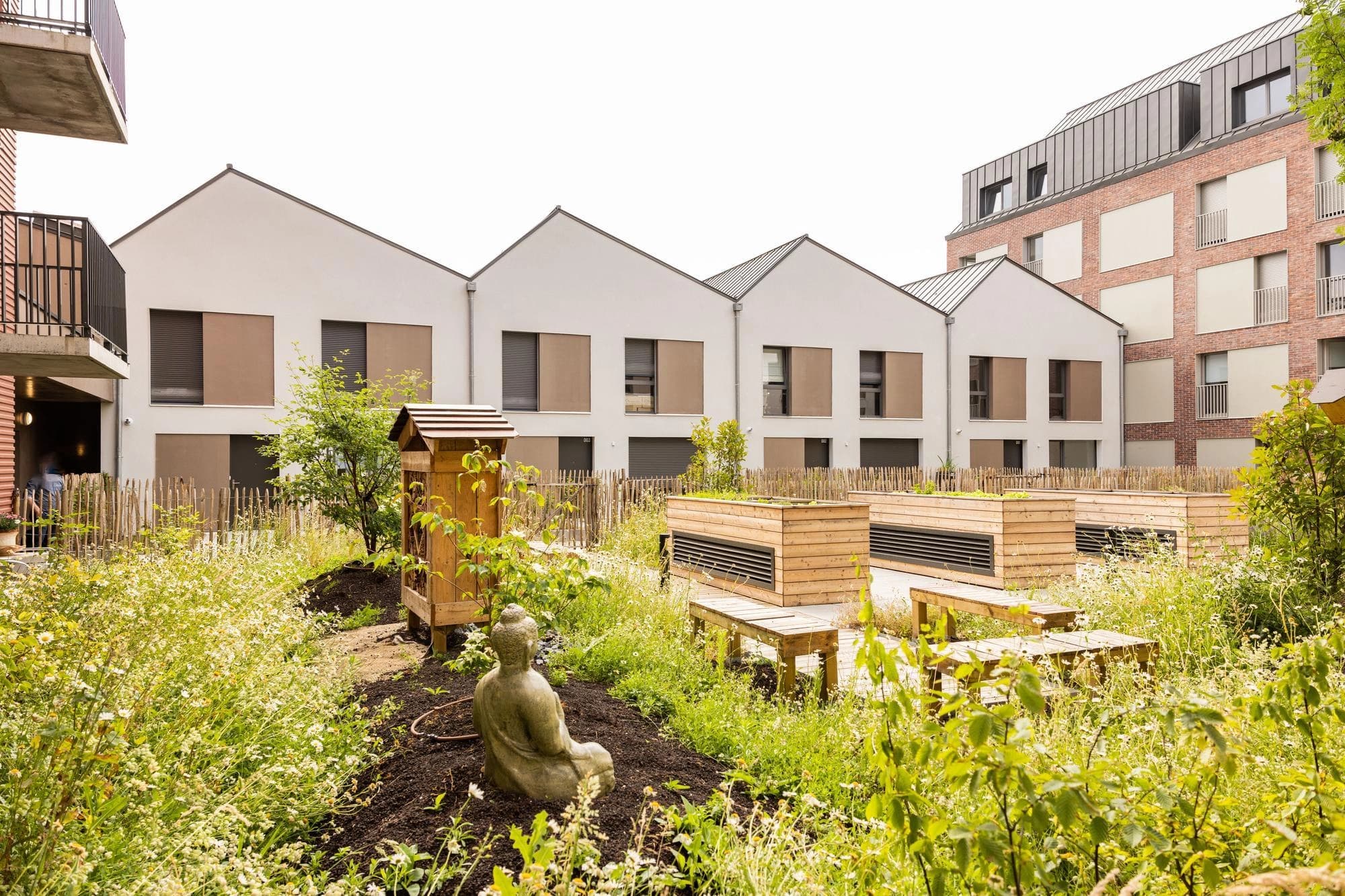 Un jardin urbain paisible avec une statue de Bouddha, des bancs en bois et des jardinières surélevées au milieu d'une végétation luxuriante et de petites fleurs blanches. En arrière-plan se trouvent des maisons modernes aux couleurs claires avec des toits sombres et un immeuble d'appartements en briques.