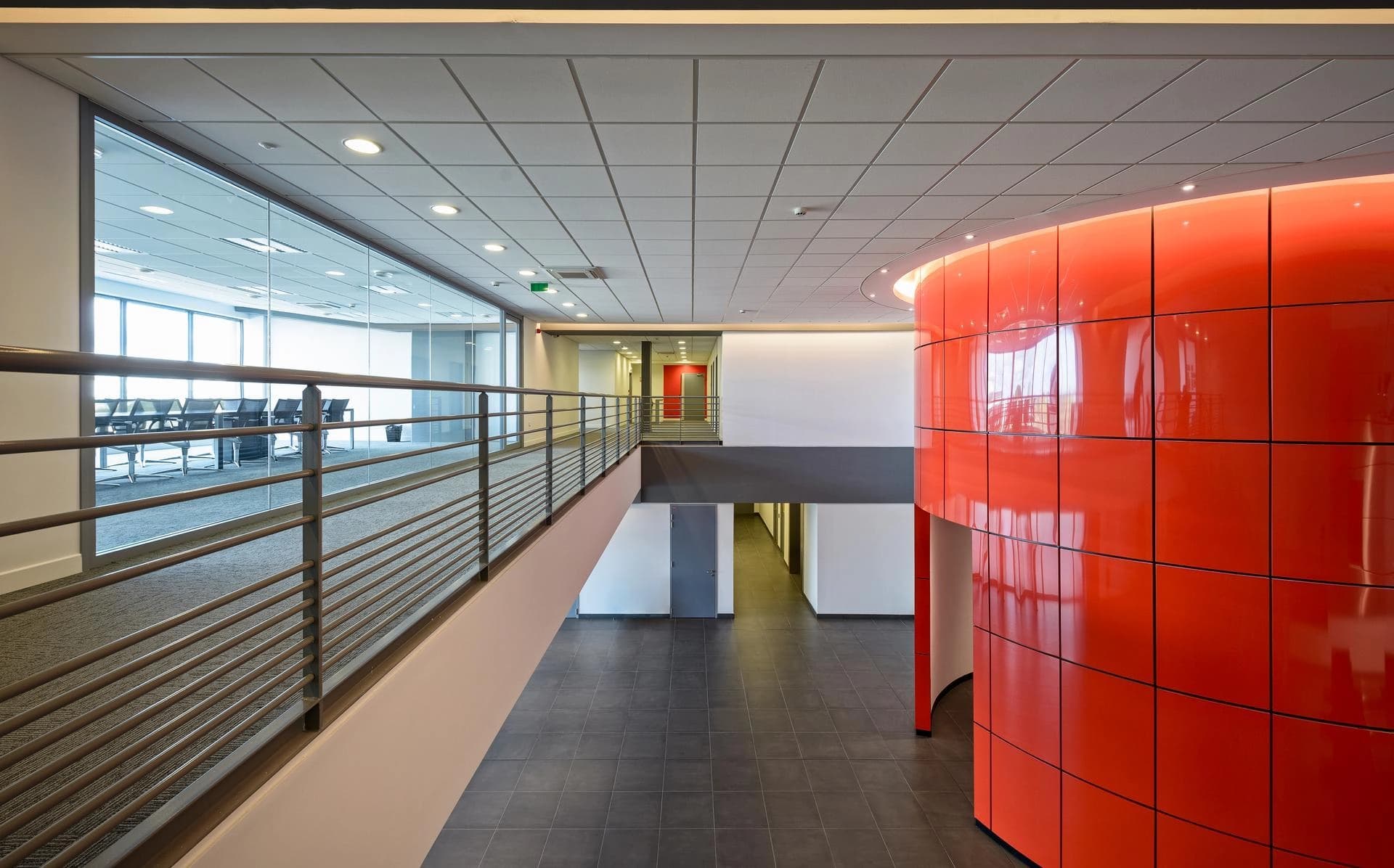Hall d'entrée moderne avec mezzanine donnant sur un rez-de-chaussée carrelé, avec un mur incurvé orange brillant à droite et des salles de conférence aux parois vitrées visibles à travers une balustrade métallique à gauche.