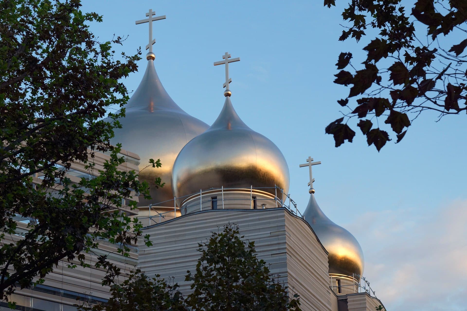 Quatre dômes d'église orthodoxe brillants et argentés, reflétant le soleil avec des teintes dorées et surmontés de croix, se dressent sous un ciel bleu clair, encadrés par des branches d'arbres.