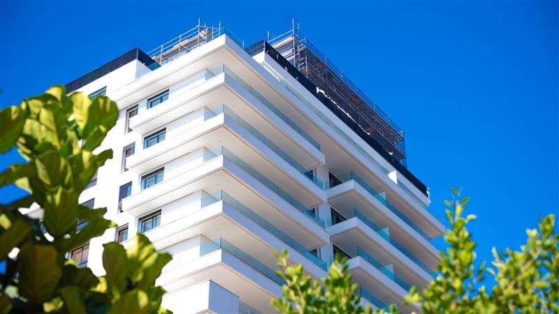 Immeuble d'habitation moderne blanc aux nombreux balcons sous un ciel bleu, entouré de feuilles vertes.