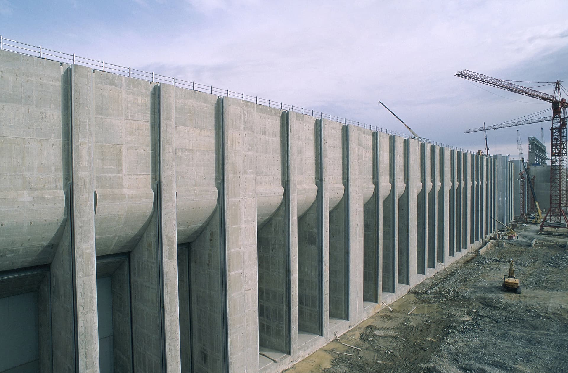 Un immense mur en béton gris clair, probablement un barrage ou une structure portante, est en construction, présentant des sections verticales proéminentes. Plusieurs grues rouges et de l'équipement lourd sont visibles sur le chantier sous un ciel nuageux.