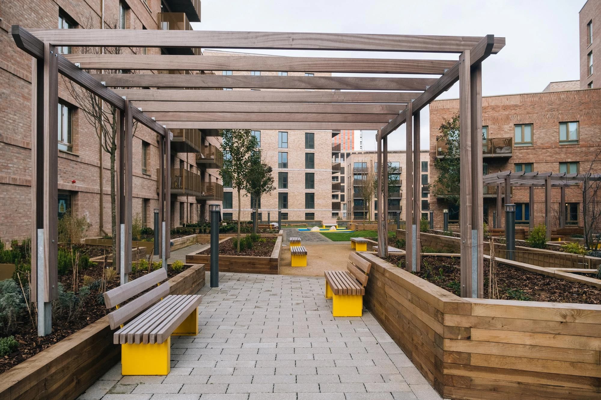 Un parvis résidentiel moderne est traversé par une pergola en bois, bordé de parterres surélevés et de bancs en bois avec des pieds jaunes vifs sur des pavés gris, le tout entouré d'immeubles en briques.