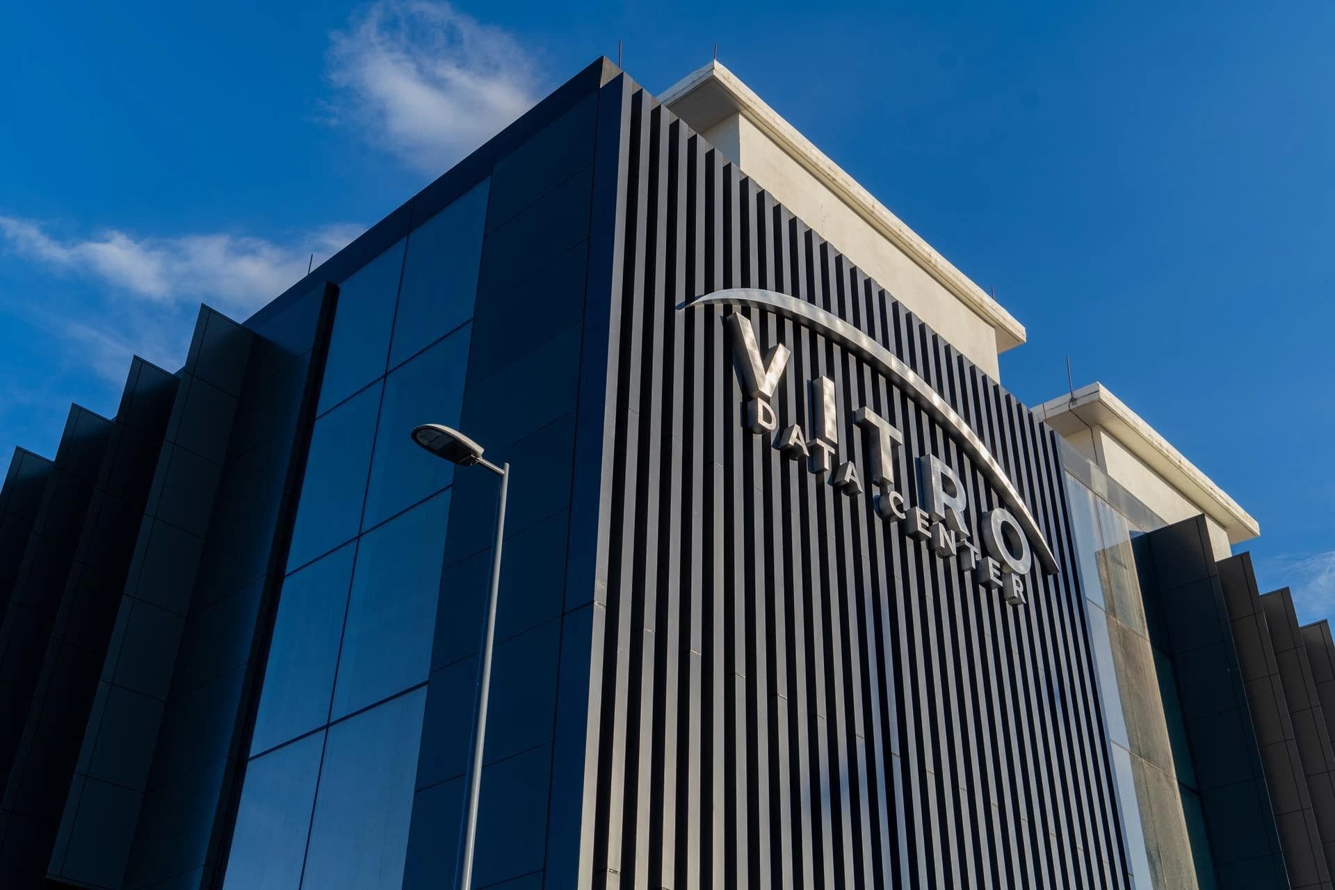 Un bâtiment moderne abritant le "VITRO DATA CENTER" présente des façades en verre sombre et des panneaux verticaux noirs, avec son logo argenté proéminent. Il se dresse sous un ciel bleu clair et ensoleillé, avec quelques nuages blancs et un lampadaire visible sur la gauche.