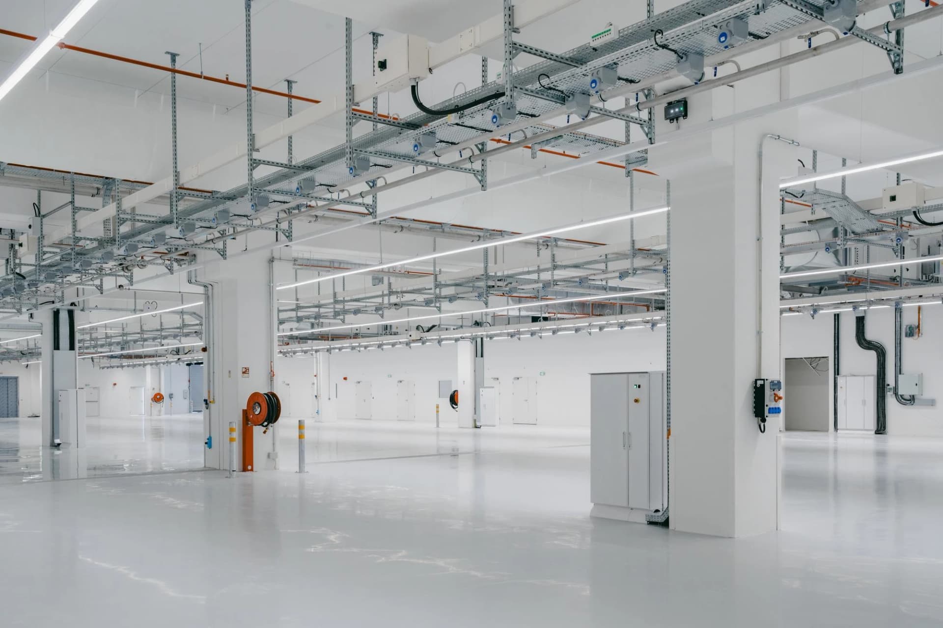 Un espace industriel moderne, vide et très lumineux, se caractérise par un sol blanc réfléchissant, des murs blancs, de vastes chemins de câbles argentés suspendus équipés de luminaires LED linéaires, ainsi que des équipements industriels visibles, comme un dévidoir de tuyau d'incendie orange.