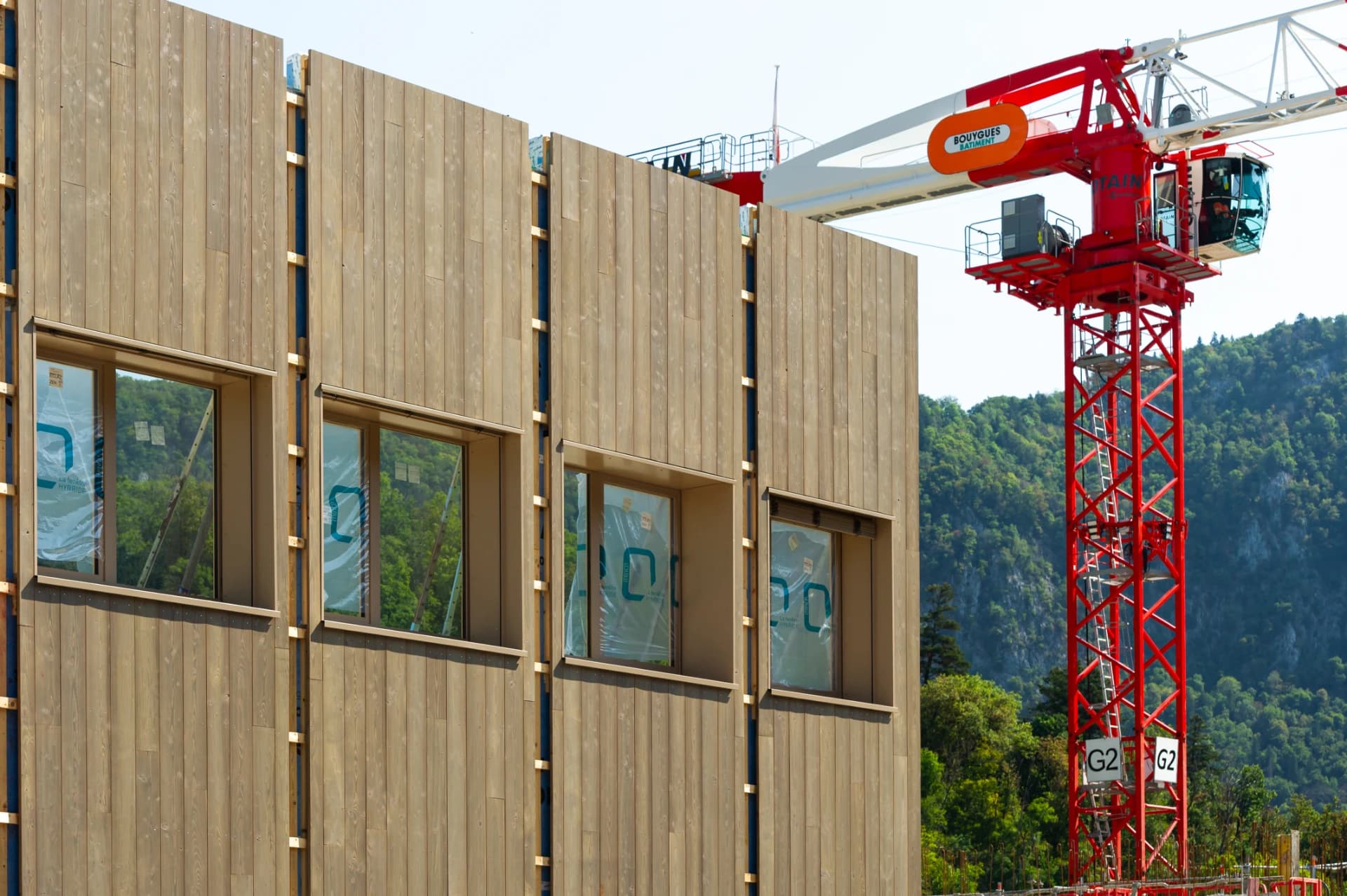 Un bâtiment en construction avec une façade en bois clair et des fenêtres, flanqué d'une grande grue rouge et blanche "BOUYGUES BATIMENT", sur fond de montagnes verdoyantes et d'un ciel clair.