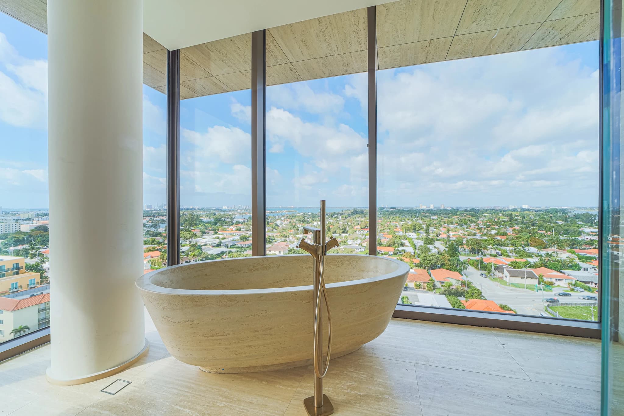 Luxueuse baignoire autoportante en travertin avec un robinet doré moderne, placée devant une fenêtre pleine hauteur offrant une vue panoramique sur une ville verdoyante et l'océan sous un ciel bleu nuageux.