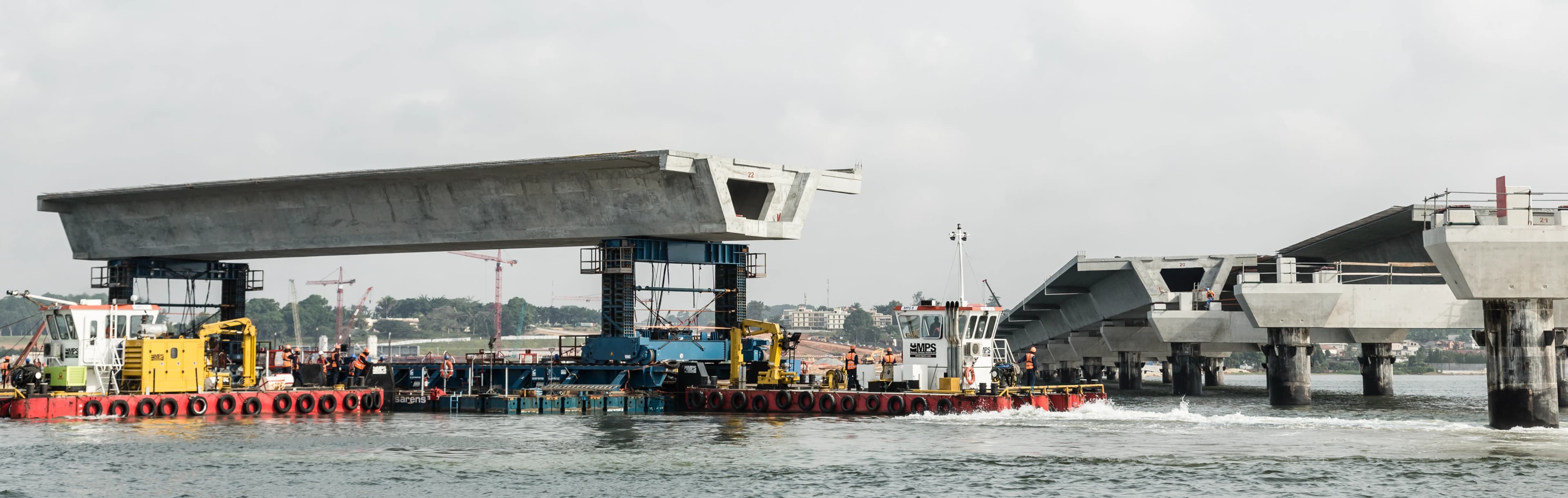 Des segments de pont en béton gris sont installés sur l'eau, avec des barges de construction, dont une avec des machines jaunes et des ouvriers en gilets orange, déplaçant un segment vers une partie du pont déjà érigée sous un ciel couvert.