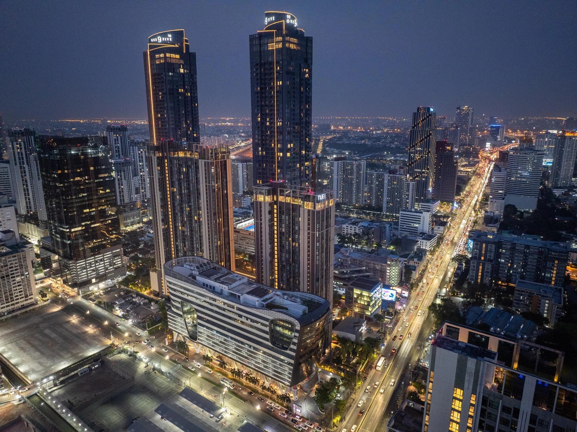 Vue aérienne nocturne de Bangkok avec des gratte-ciel illuminés et une autoroute aux lumières scintillantes.