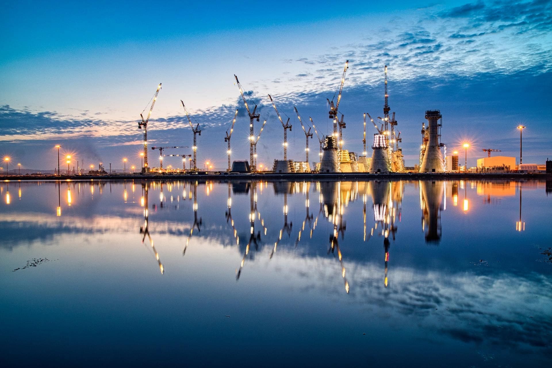 Vaste chantier industriel nocturne avec de nombreuses grues et de grandes structures en béton illuminées, dont les reflets orange vif animent l'eau calme et sombre sous un ciel bleu-gris nuageux.