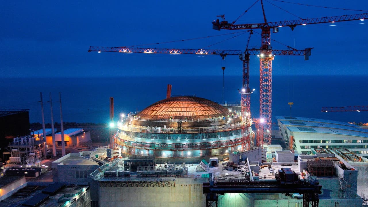 Chantier de construction nocturne éclairé, dominé par un grand bâtiment de réacteur à dôme orange avec échafaudages, entouré de hautes grues rouges. Le complexe industriel est situé au bord d'une mer bleu foncé sous un ciel sombre.