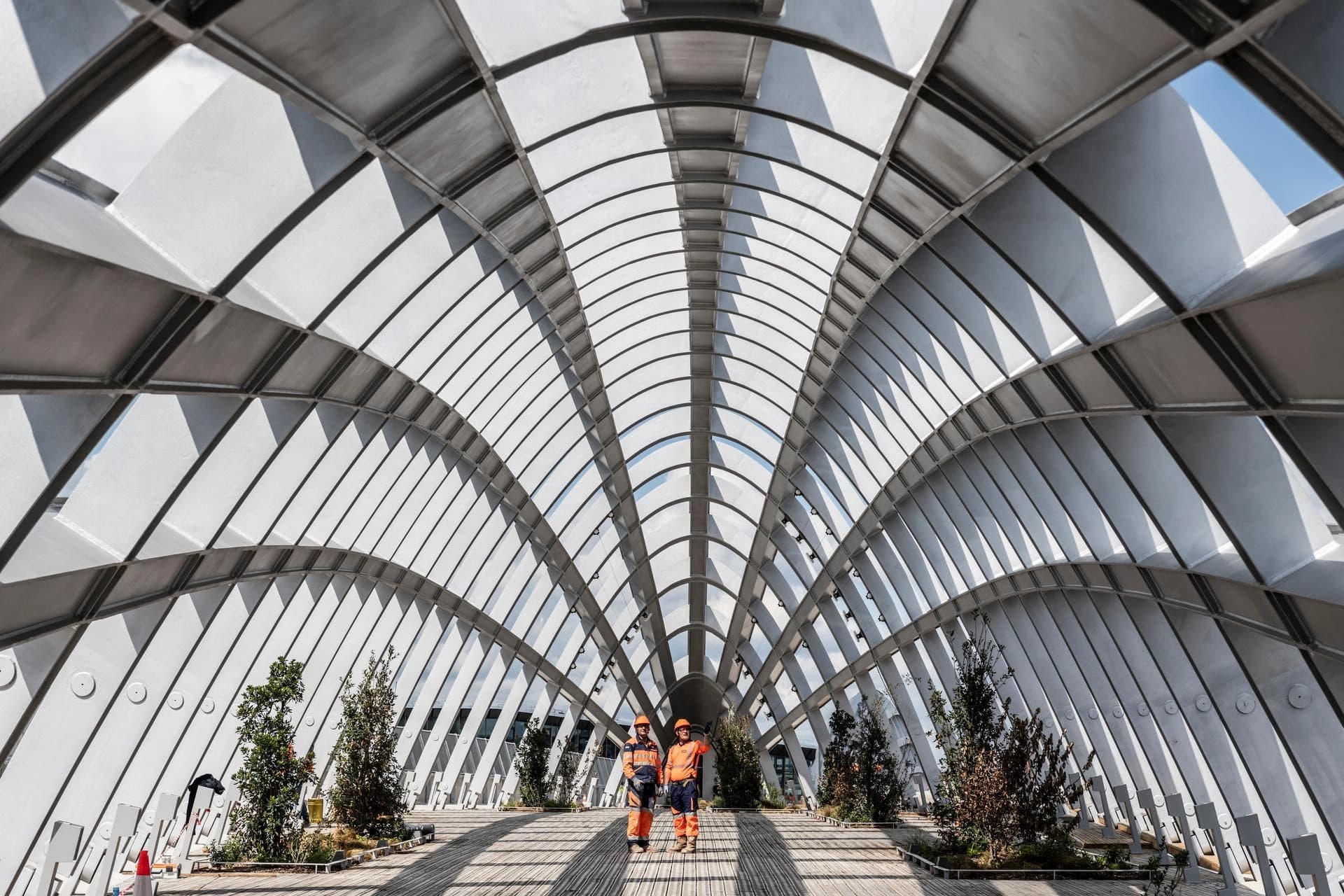 Deux ouvriers en tenues de sécurité orange et bleues avec casques se tiennent sur un plancher en bois sous un immense toit architectural incurvé fait de larges poutres métalliques grises et de panneaux translucides, baigné de lumière du soleil.
