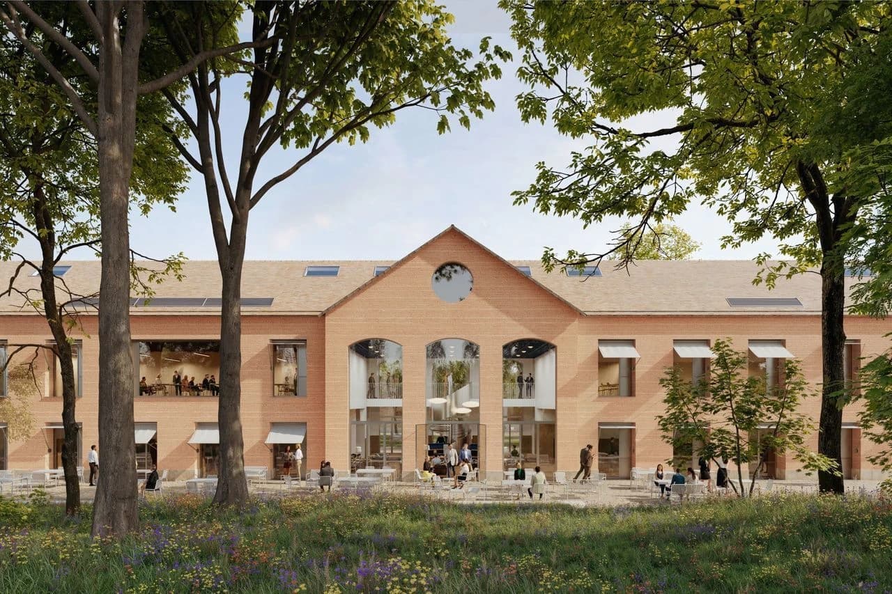 Grand bâtiment moderne en briques orange avec un toit clair, de larges fenêtres et un fronton central orné d'une fenêtre ronde. Des personnes sont assises sur une terrasse extérieure meublée de tables blanches, devant un jardin fleuri et encadré par de grands arbres.