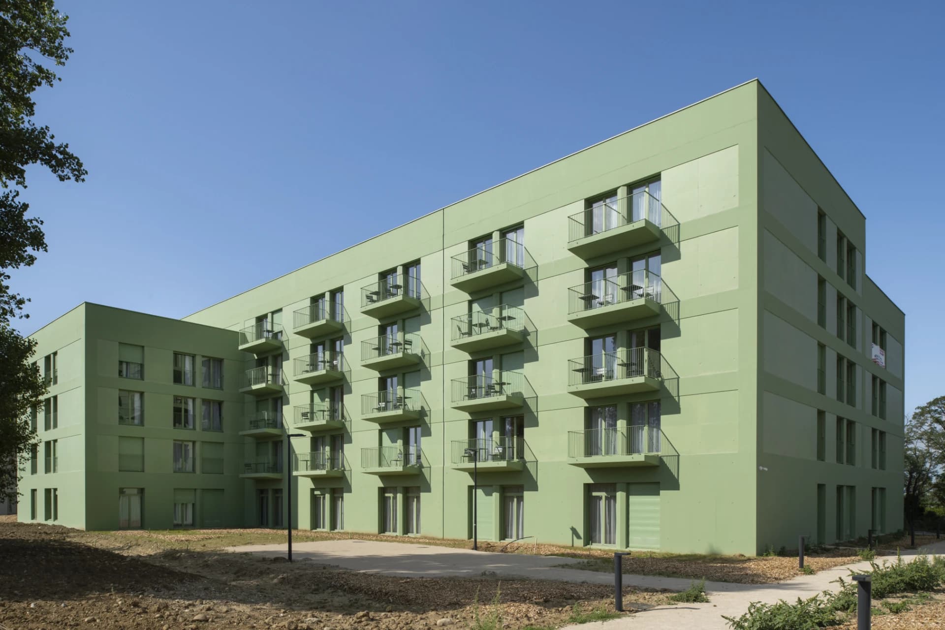 Un bâtiment résidentiel moderne de plusieurs étages, aux façades vert pastel et multiples balcons avec tables et chaises, est vu sous un ciel bleu clair. Le terrain devant est partiellement aménagé, avec un panneau "À LOUER" visible sur l'une des façades.