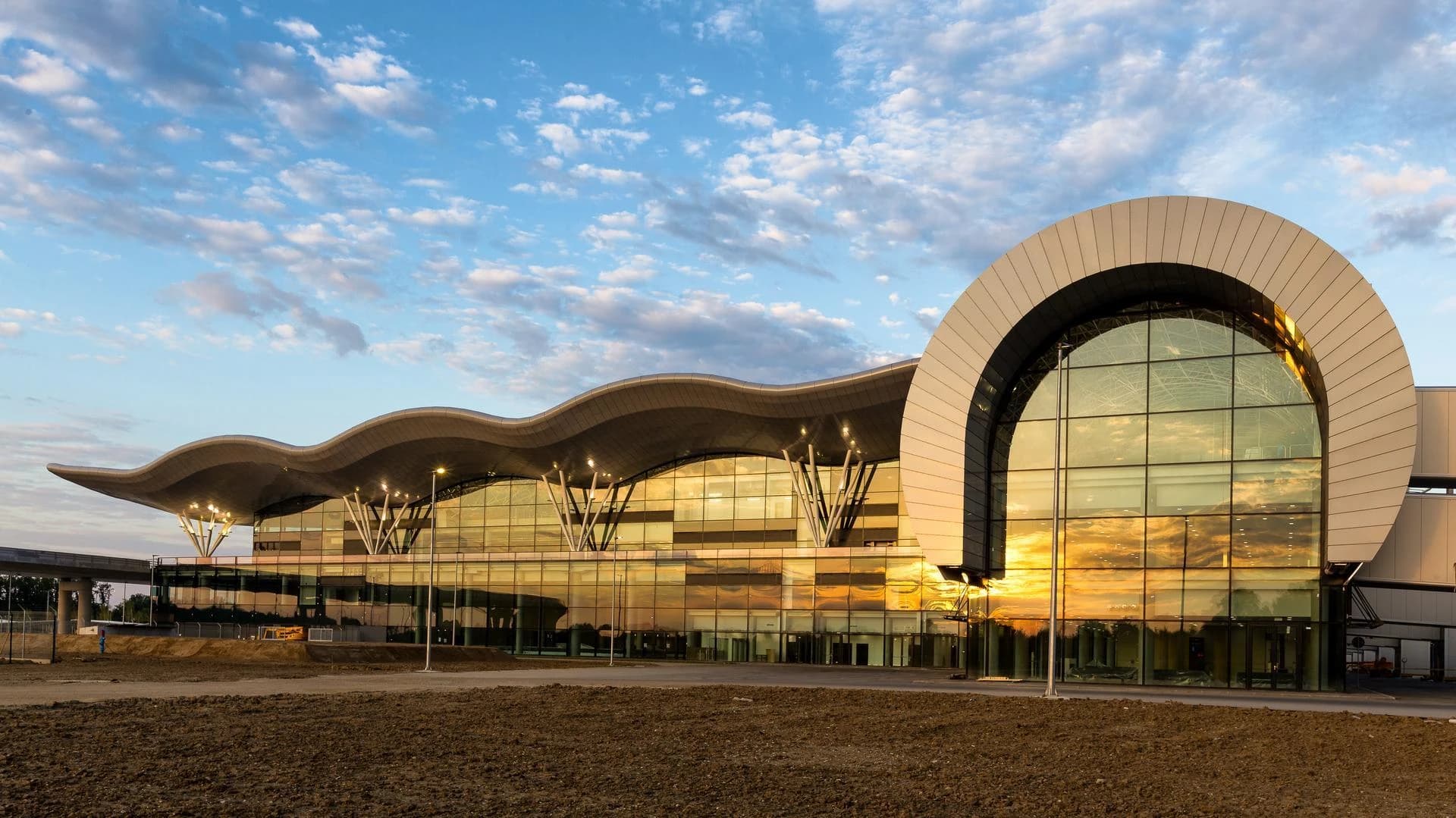 Terminal d'aéroport moderne au toit ondulé et à la façade vitrée réfléchissant un ciel bleu nuageux et un coucher de soleil doré, avec un sol en terre au premier plan.