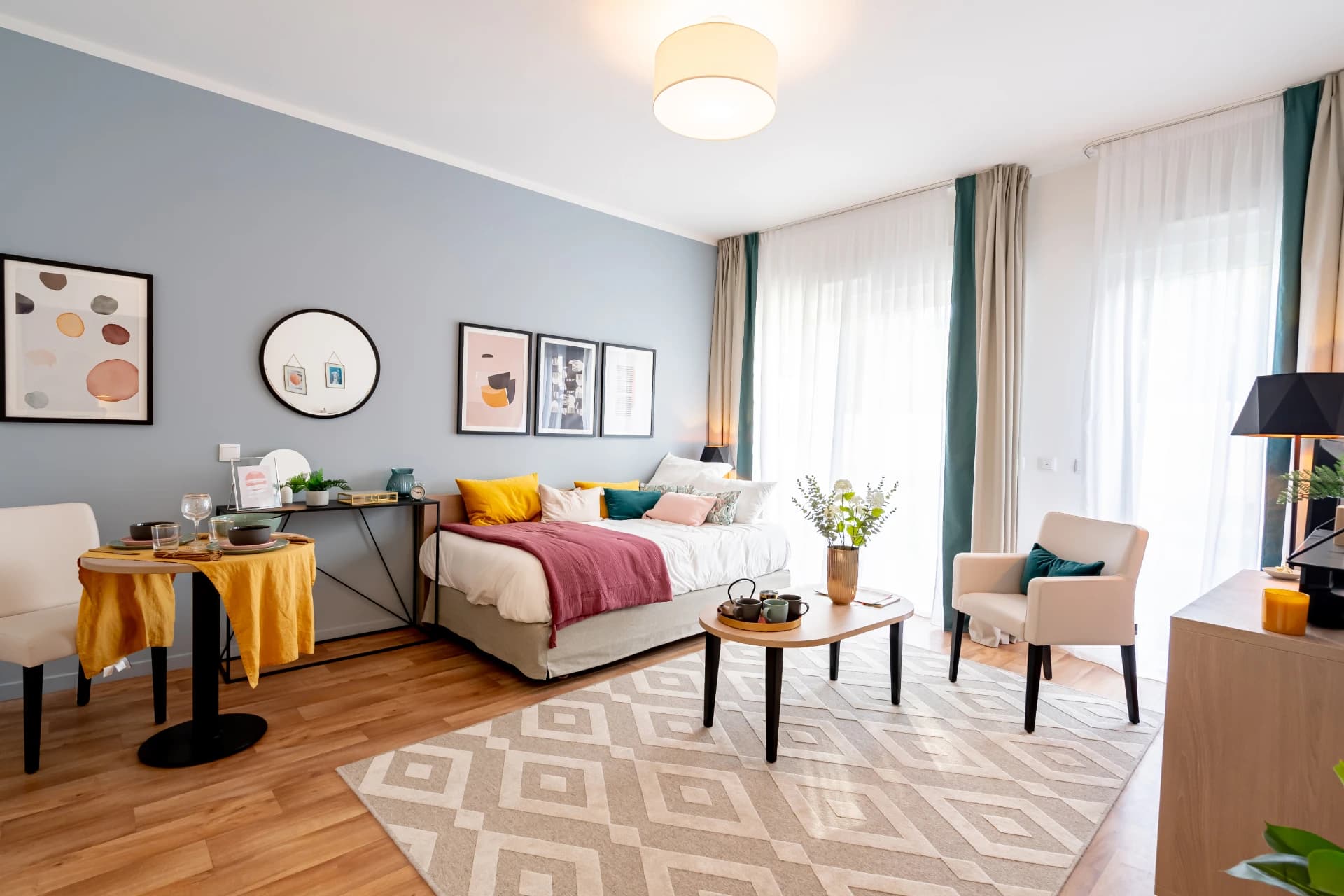 A bright, modern studio apartment features a bed with vibrant pillows, a small dining area, and an armchair on a geometric rug, all against light blue walls with framed art.