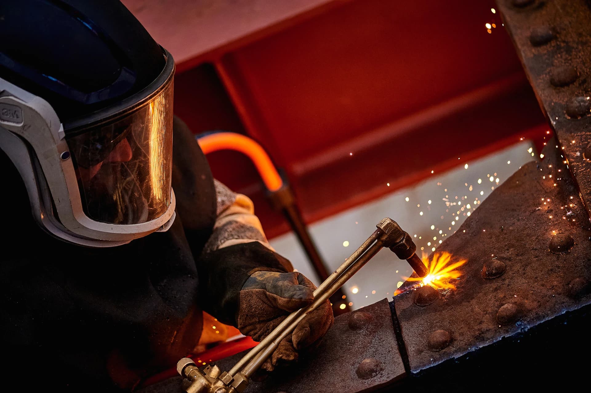 Un ouvrier portant un casque de protection sombre et des gants utilise un chalumeau pour couper une pièce de métal rouillée, créant de vives étincelles jaunes et orange. La scène sombre est éclairée par la lumière intense de l'opération, avec des structures métalliques rouges en arrière-plan.