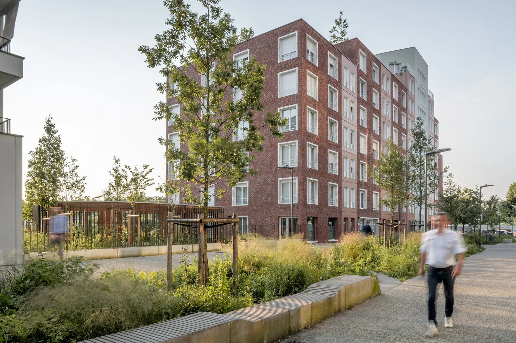 Un bâtiment moderne de plusieurs étages, avec des façades en briques rouge-brun et des sections beige clair, présente de nombreuses fenêtres encadrées de blanc au milieu d'arbres verdoyants et de parterres de jardin. Des silhouettes floues de personnes marchent sur un chemin pavé, soulignant l'aménagement paysager urbain animé.