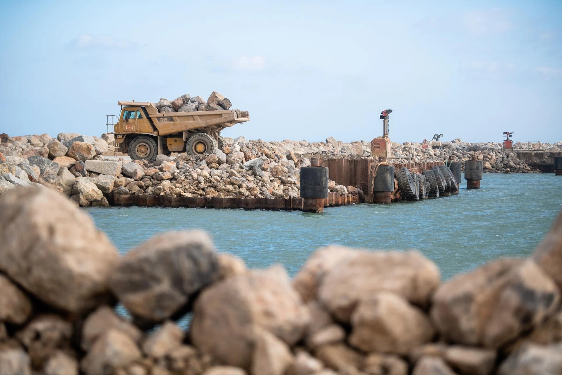 Un camion benne jaune CAT rempli de roches se trouve sur une digue rocheuse, avec des structures de quai rouillées et des balises rouges longeant l'eau bleue sous un ciel clair.