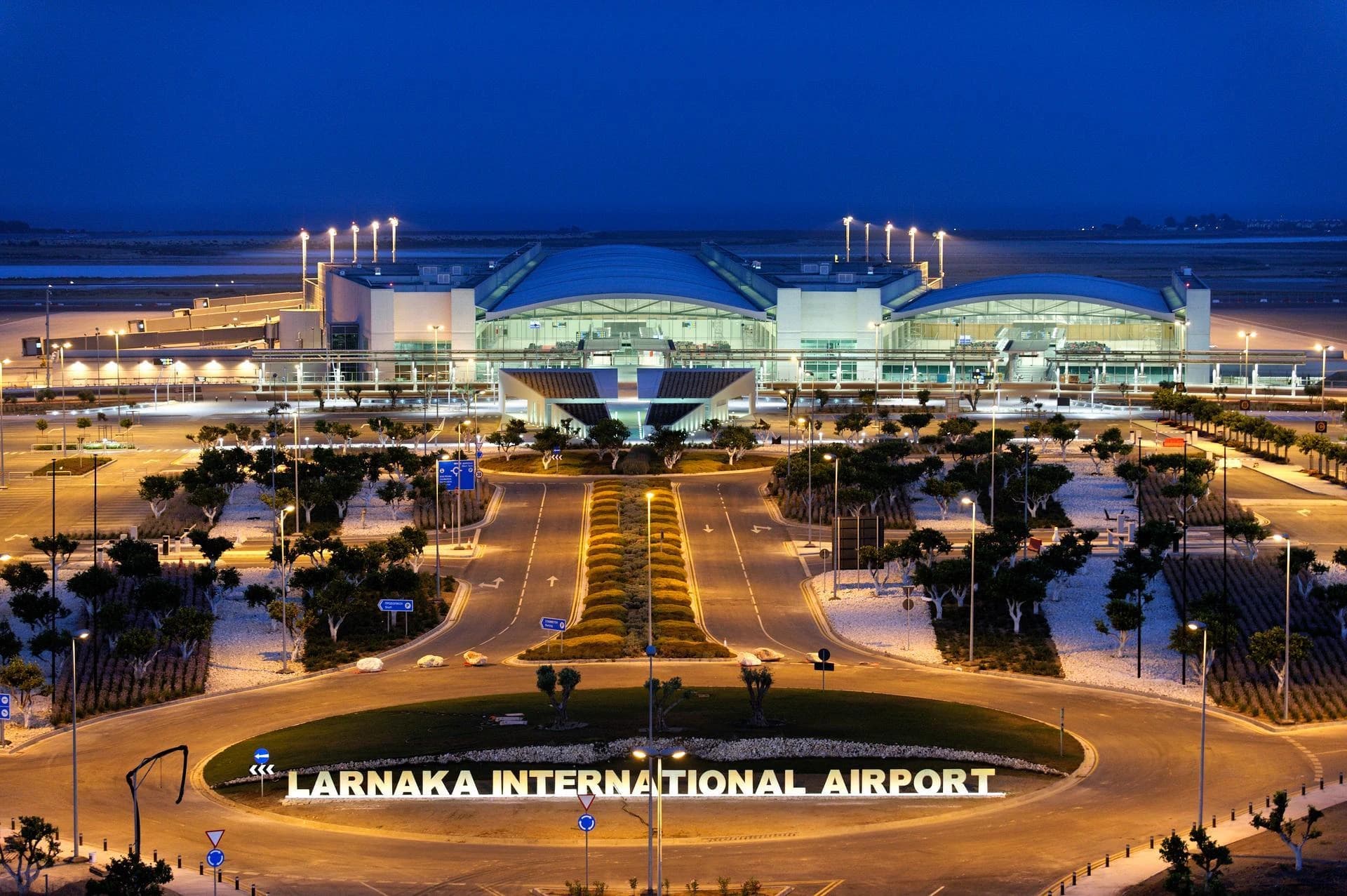 Vue nocturne de l'aéroport international de Larnaka, montrant le terminal moderne illuminé, une grande enseigne "LARNAKA INTERNATIONAL AIRPORT" au premier plan, et un réseau de routes et d'aménagements paysagers éclairés sous un ciel bleu foncé.