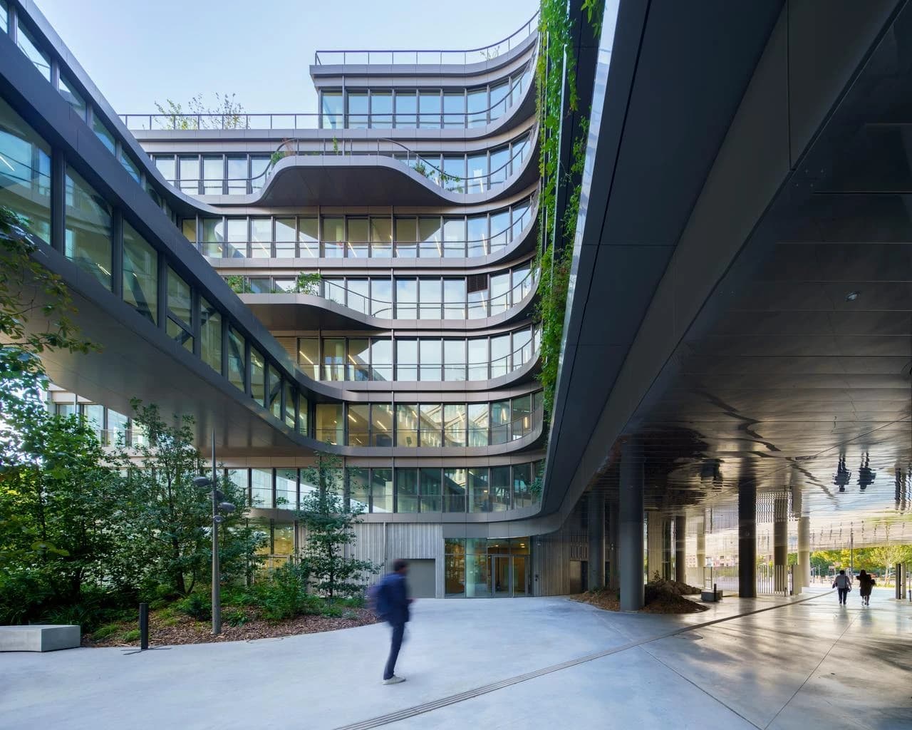 Un immeuble moderne à la façade ondulante en verre et balcons végétalisés, avec un jardin vertical luxuriant. Un pont piéton courbe le relie à une place aménagée où des personnes marchent sous un ciel clair.