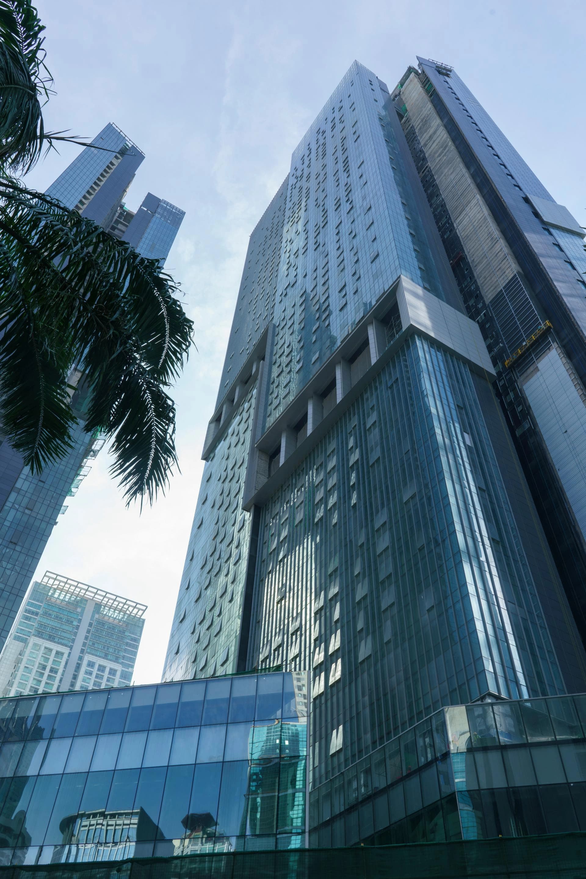 Plusieurs gratte-ciel modernes aux façades de verre bleu-vert s'élancent vers un ciel clair et lumineux, encadrés par des feuilles de palmier vert foncé en premier plan.