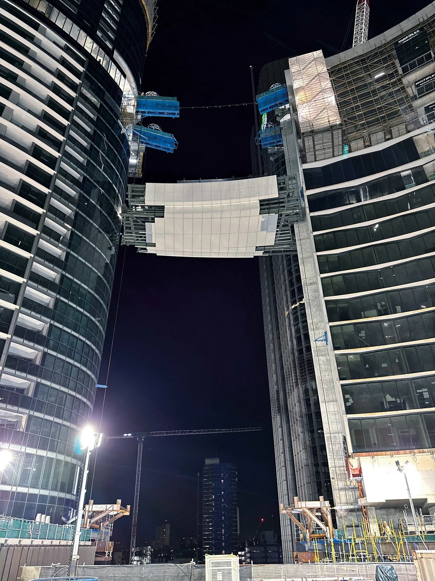 Vue nocturne de deux tours modernes en construction, aux façades de verre sombre et béton clair, reliées par une passerelle aérienne. Des équipements de chantier, dont des grues et des plateformes suspendues bleues, illuminent les structures sous un ciel noir.