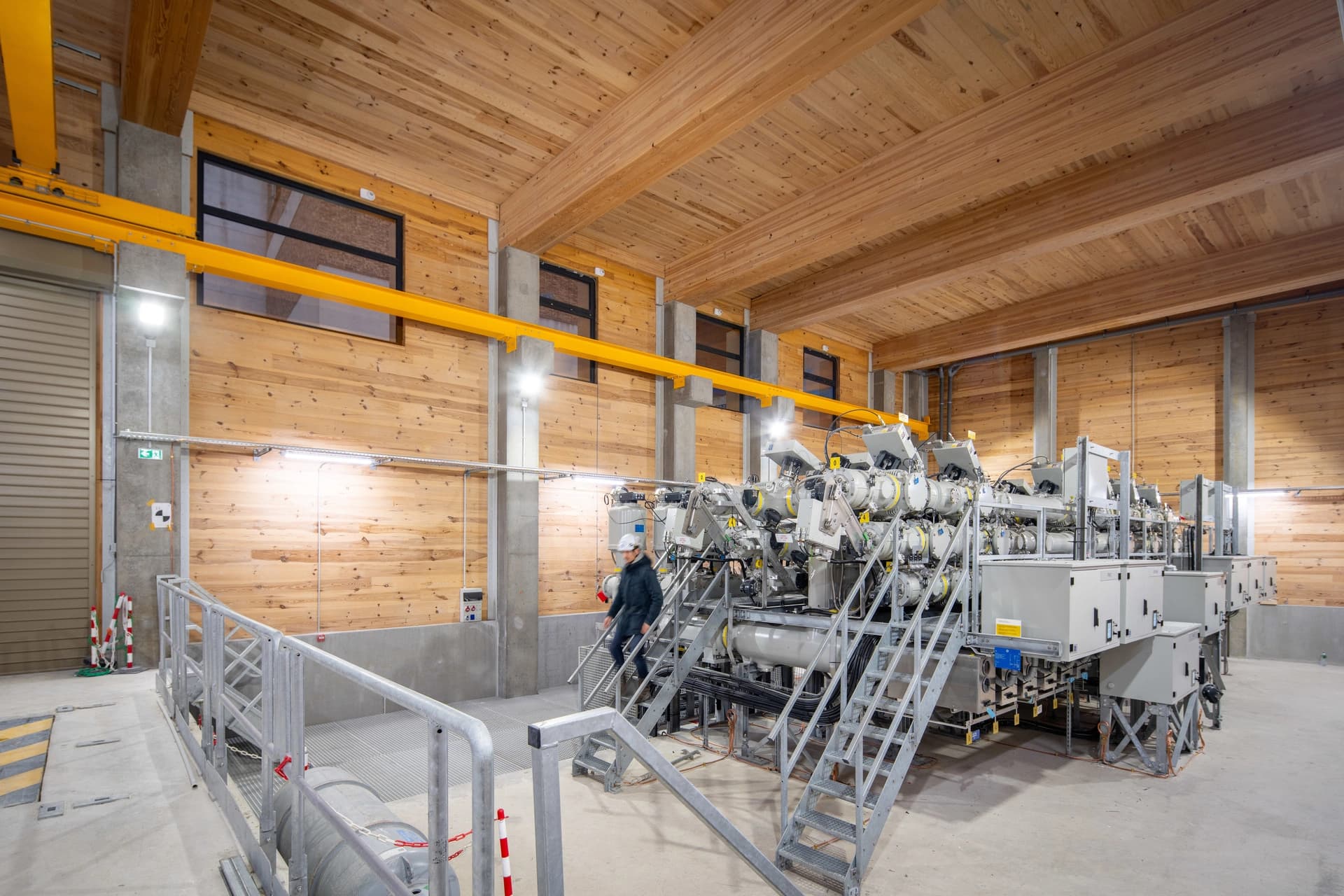 Intérieur d'un bâtiment industriel moderne avec des murs en bois clair et des poutres apparentes au plafond, abritant de grandes machines électriques grises et blanches dotées d'escaliers métalliques. Un travailleur monte ces escaliers, sous une poutre de grue jaune.