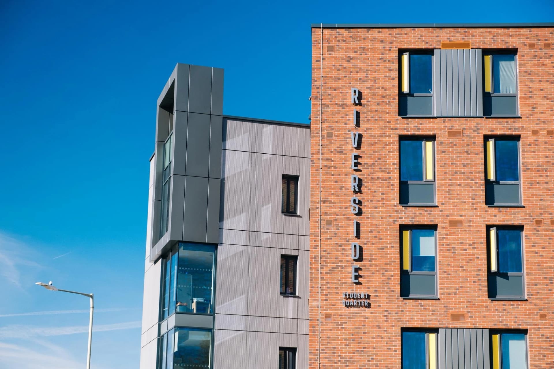 Façades de deux bâtiments modernes sous un ciel bleu clair : l'un en briques orange-rouge avec l'inscription verticale "RIVERSIDE STUDENT QUARTER" et des fenêtres aux reflets bleus et jaunes, l'autre en panneaux gris et verre.