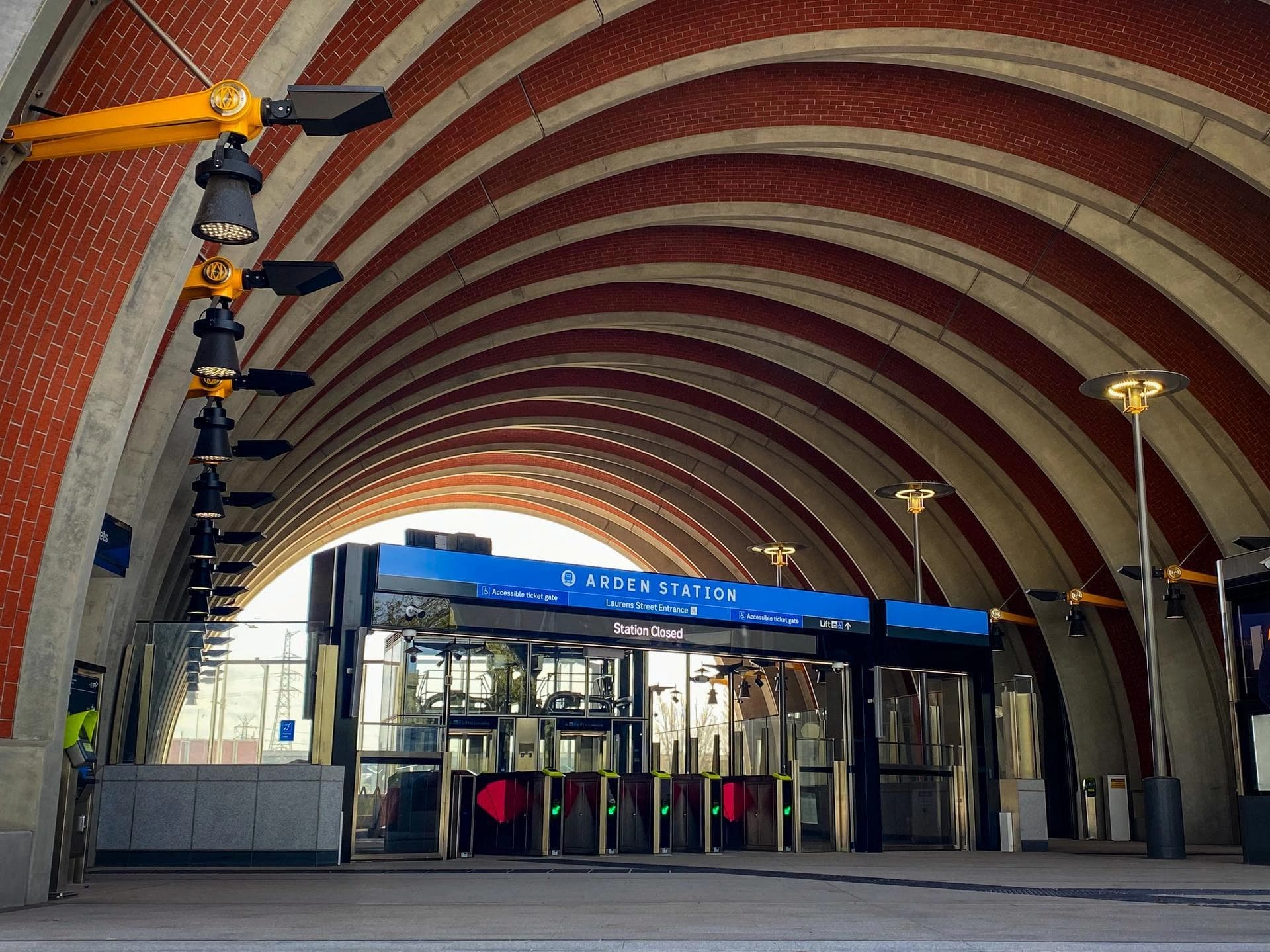 Un large plan de l'entrée moderne de la station Arden, présentant un plafond voûté avec des arches alternant briques rouges et béton gris. En dessous, le panneau "Station Closed" est visible au-dessus de portiques de billetterie, éclairé par des luminaires industriels jaunes et noirs.
