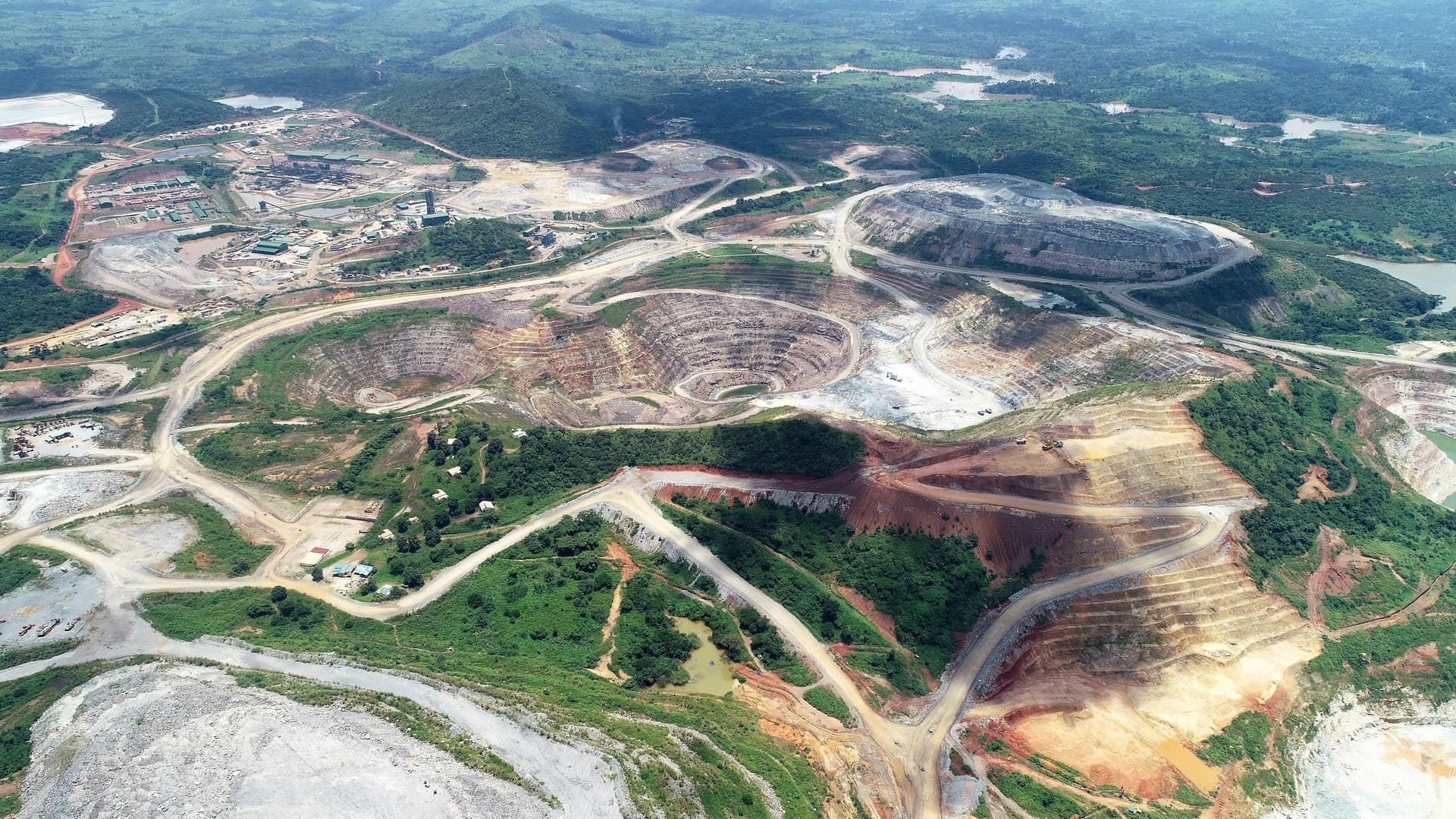 Vue aérienne d'un vaste complexe minier à ciel ouvert présentant de multiples fosses terrassées aux couleurs variées (terre, gris, blanc), un réseau routier étendu et des infrastructures industrielles, s'étendant au milieu de collines verdoyantes et de plans d'eau.