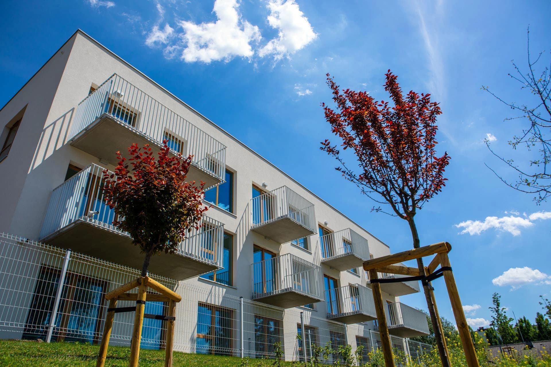 Un immeuble résidentiel blanc moderne avec des balcons et des arbres rouges sous un ciel bleu ensoleillé.