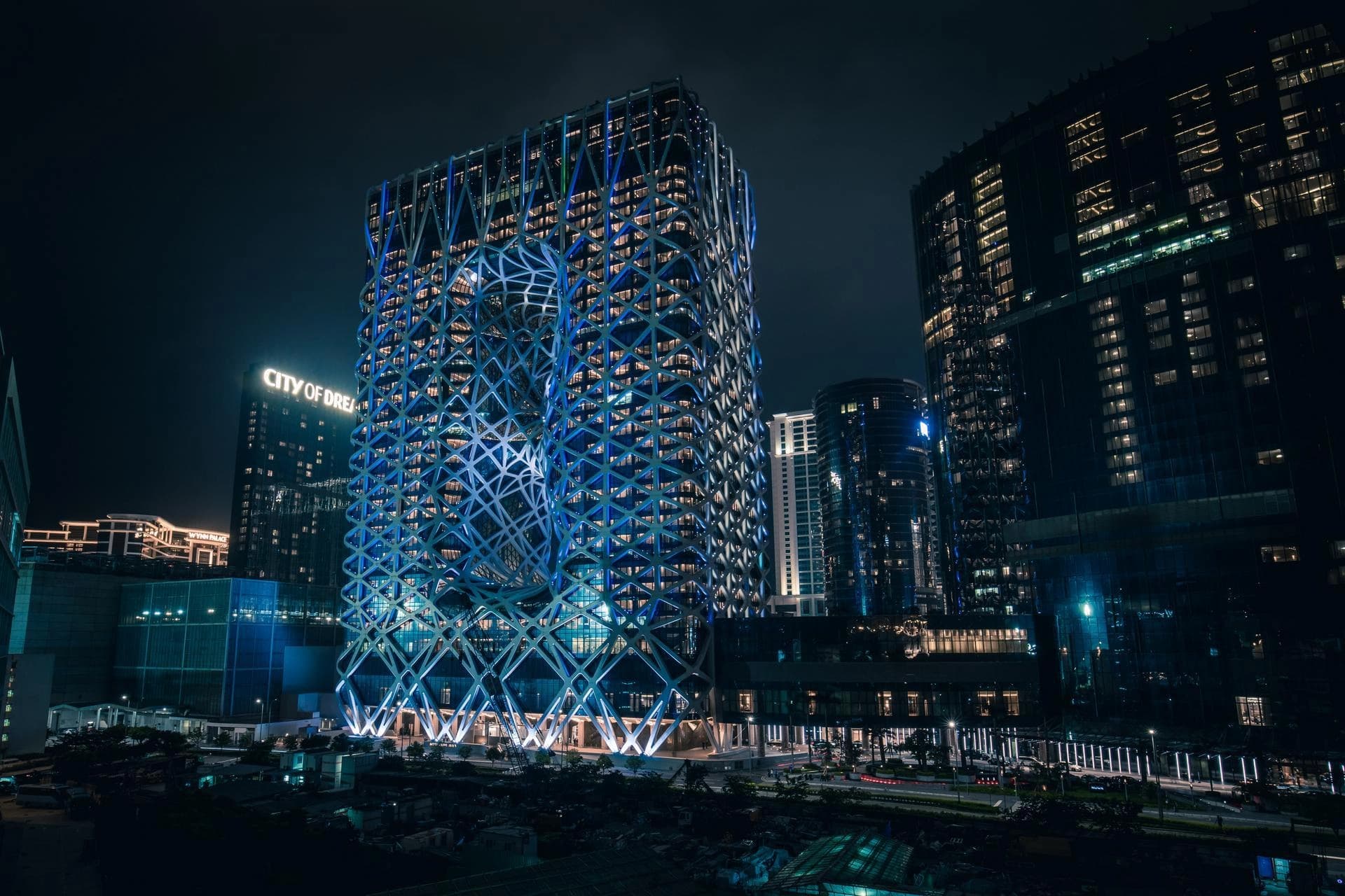 Un gratte-ciel moderne et futuriste, le Morpheus Hotel, illumine la nuit avec sa façade en treillis géométrique complexe et ses ouvertures sculpturales, brillamment éclairée en bleu. D'autres tours sombres aux fenêtres jaunes s'élèvent autour dans ce paysage urbain nocturne.