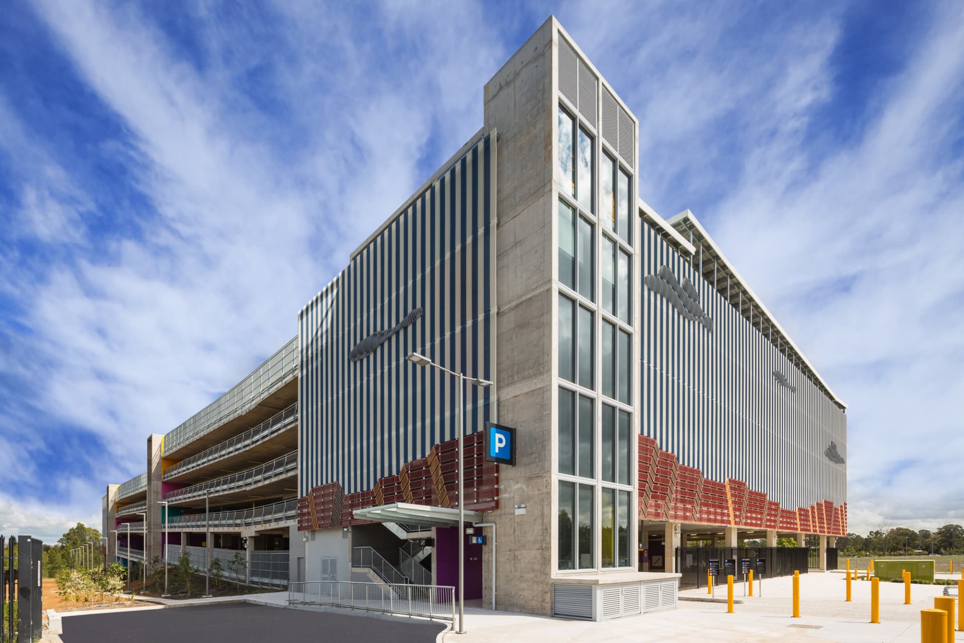 Parking moderne à plusieurs niveaux avec une façade rayée, une cage d'escalier vitrée et un ciel bleu.