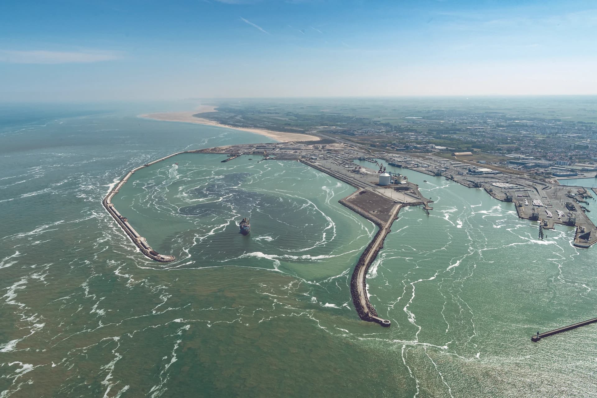 Vue aérienne d'un vaste port maritime avec ses longs brise-lames protégeant des navires et des infrastructures industrielles, longeant une côte sablonneuse et une ville sous un ciel bleu clair. L'eau verte et bleue présente des tourbillons d'écume blanche, s'étendant vers l'océan lointain.