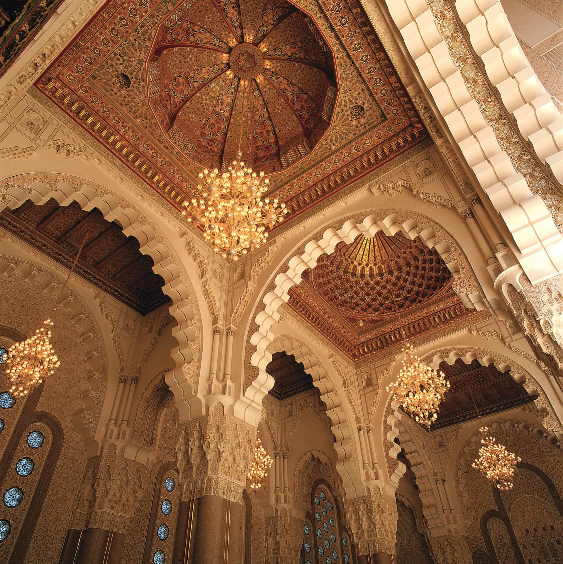 L'intérieur somptueux d'une mosquée marocaine présente des plafonds et dômes richement décorés de motifs géométriques et floraux rouges, or et bruns, illuminés par de grands lustres et des arches sculptées crèmes.